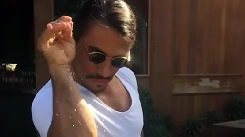 Salt Bae — Chef Nusret Gökçe captured mid-gesture performing signature theatrical salt sprinkle in white tank top and sunglasses.