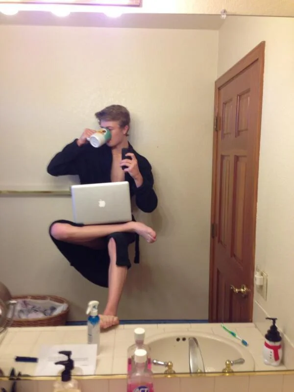 A creative Selfie Olympics variant showing a person balanced on a laptop while taking a bathroom mirror self-portrait.