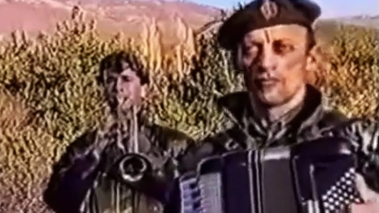 Serbia Strong — Man in military beret playing accordion outdoors with brass player behind him, still from Serbian nationalist music video.