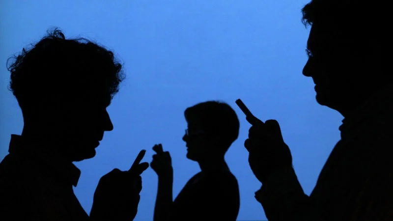 Shadow Banning Shadowban — Three dark human silhouettes against a vivid blue background, each holding a small device, creating a shadowy aesthetic evoking