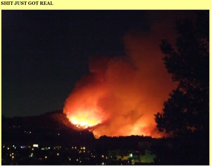 Nighttime wildfire burning across hills above a city with 'SHIT JUST GOT REAL' text
