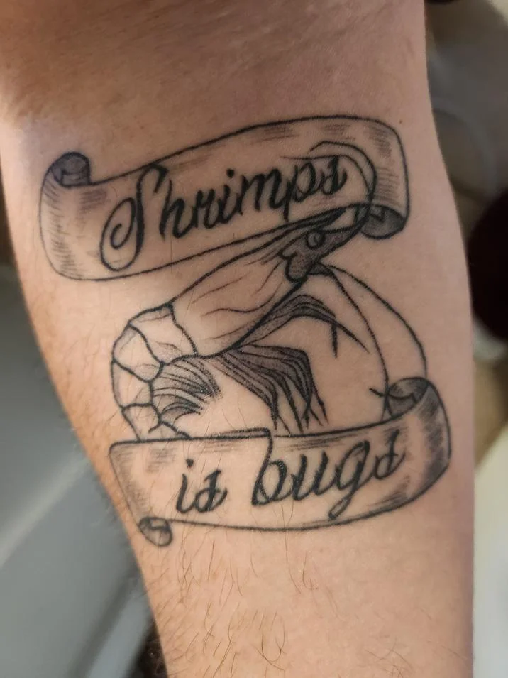Black ink forearm tattoo of a shrimp between scrolls reading 'Shrimps is bugs'