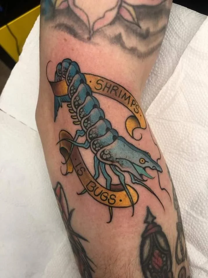Blue traditional-style shrimp tattoo on an arm with gold banners reading 'Shrimps Is Bugs'