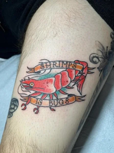 Color tattoo on a thigh showing a red shrimp framed by banners reading 'Shrimps Is Bugs'