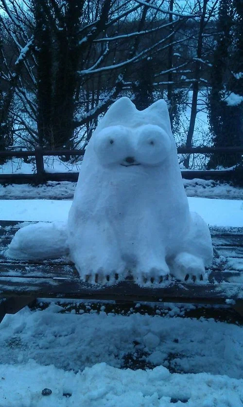 Snow sculpture of Simon's Cat with pointed ears and round eyes, sitting on a snowy wooden platform outdoors.