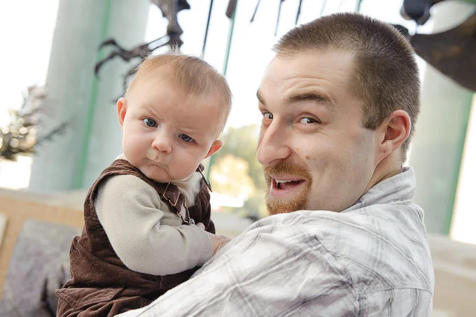 A candid photograph of the baby who became the Skeptical Baby meme, shown with an adult male figure, displaying a characteristic skeptical and questioning