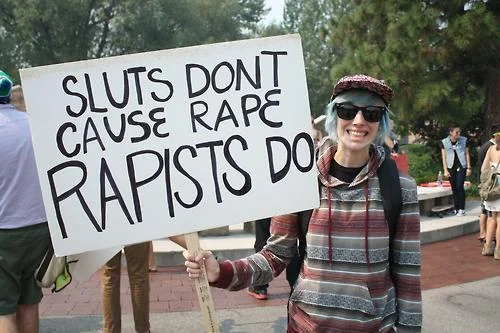 Slut Shaming — A woman with blue hair holds a large handwritten sign at an outdoor rally stating 'SLUTS DONT CAUSE RAPE RAPISTS DO'.