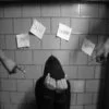 Slut Shaming — A dark, low-quality black-and-white photograph showing a person hunched against a wall with posted notes, evoking social isolation.