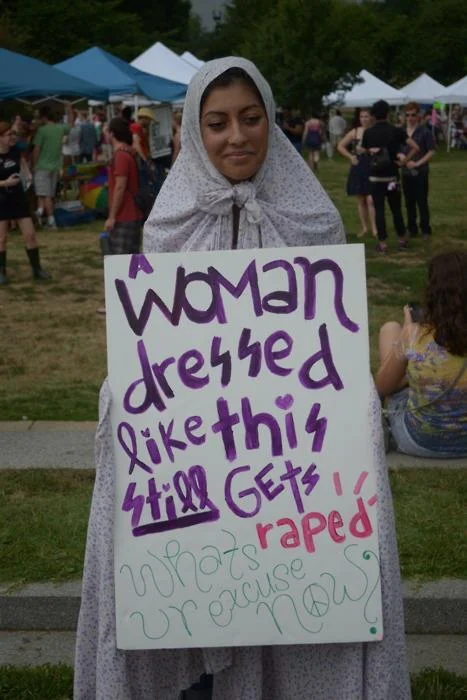 Slut Shaming — A woman wearing conservative floral headscarf holds a sign about modest dress and rape reality, dismantling shaming arguments.