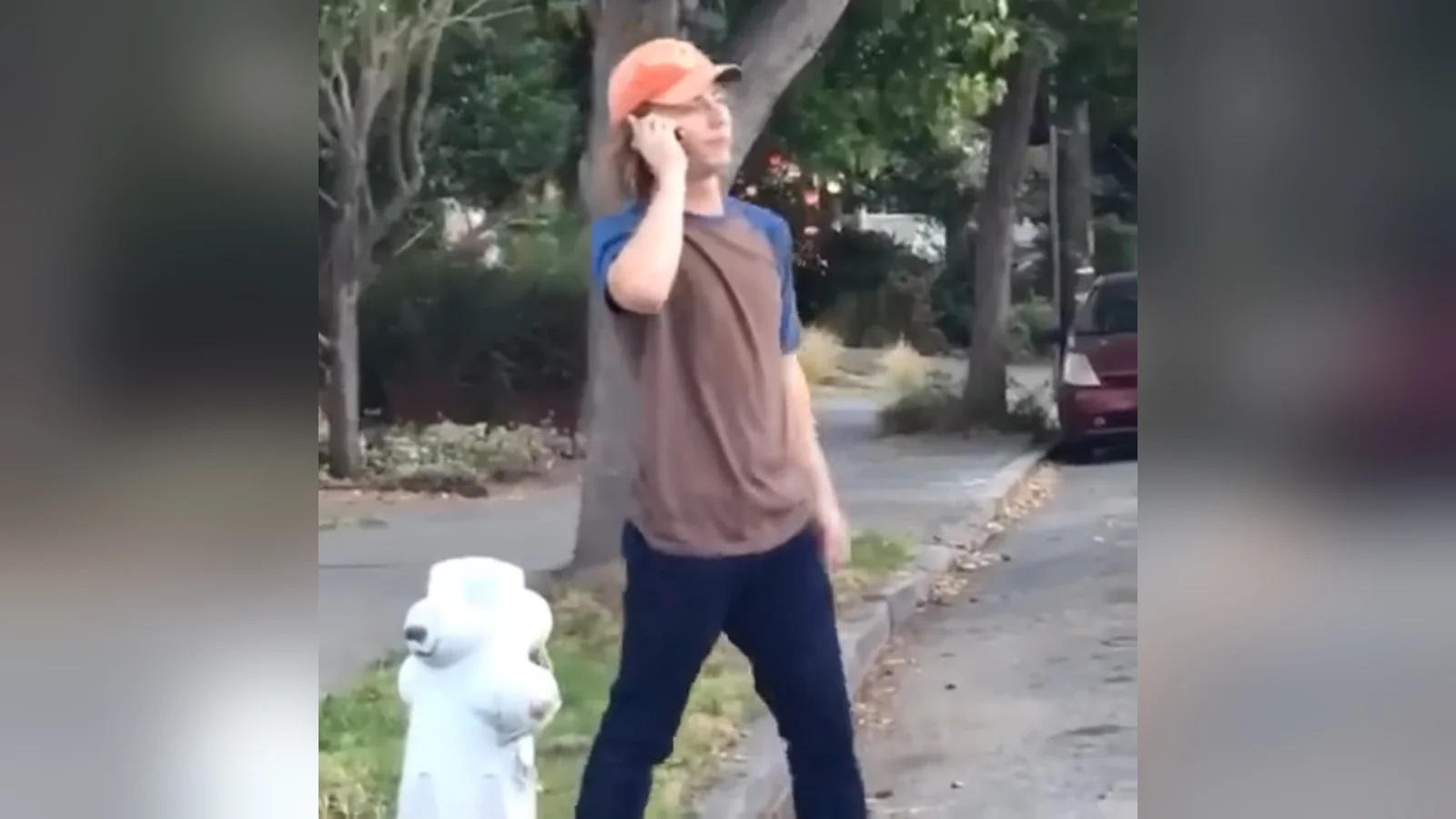 Young man in orange cap talking on a cell phone on a suburban sidewalk next to a fire hydrant, from the original 'So No Head?' Vine.