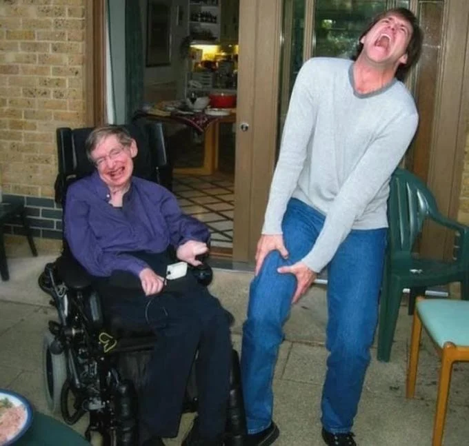 Stephen Hawking sitting in wheelchair next to a man laughing uncontrollably on a patio