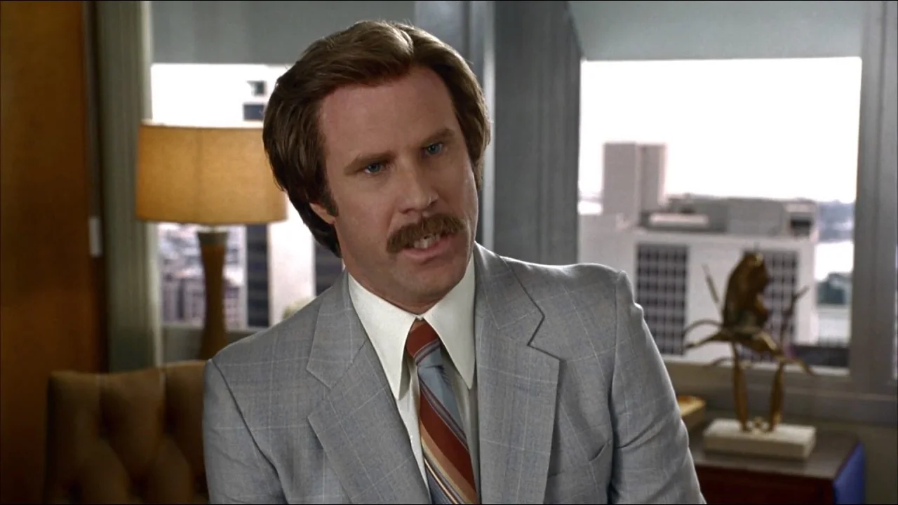Stings the Nostrils - In a Good Way — Will Ferrell as Ron Burgundy in a grey suit, mid-sentence, seated in a 1970s-styled office in Anchorman.