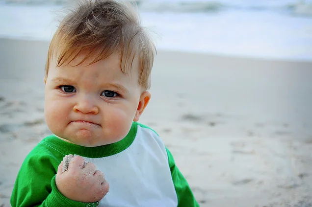 Original beach photograph of Success Kid character in green and white shirt with determined frown and clenched fist.