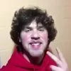 Suh Dude — Low-resolution screenshot of young man with curly dark hair in red hoodie raising two fingers.