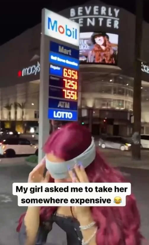 Taking My Girl Somewhere Expensive (The Gas Station) — Blindfolded red-haired woman standing in front of a Mobil gas station price sign showing high prices