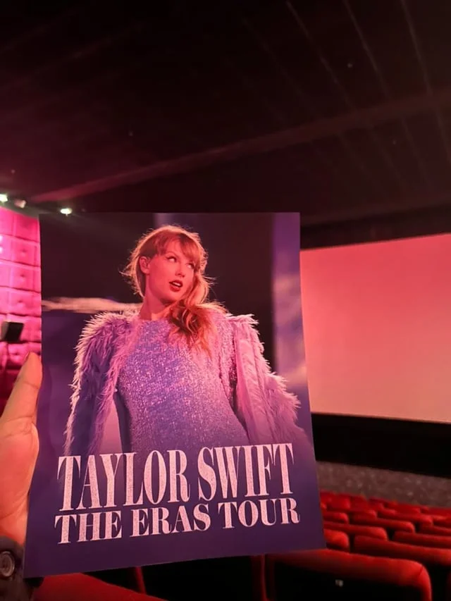 Taylor Swift Eras Tour — Fan holding Eras Tour concert film program in front of theater screen.