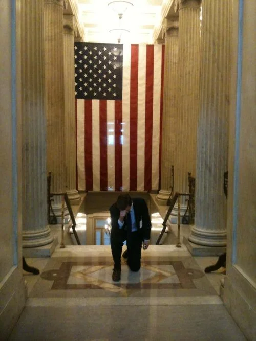 Figure performing Tebowing in classical government building interior with flag.