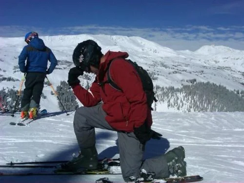 Snowboarder performing Tebowing pose on snow-covered mountain with landscape.