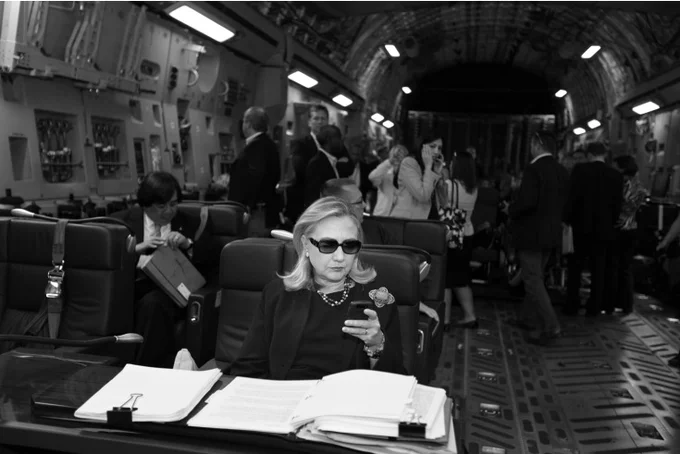 Texts From Hillary — Hillary Clinton wearing sunglasses reading a BlackBerry phone on a military plane