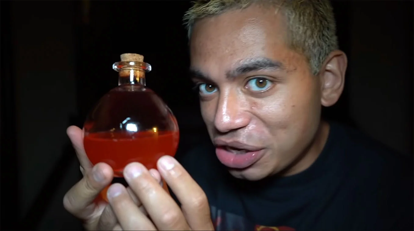 The Among Us Potion — Person holding a small cork-stopped glass bottle filled with orange-red liquid, staring at the camera in a dark room.