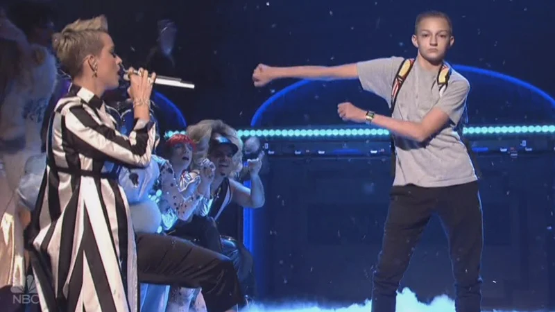 Teen boy performing the Floss dance on SNL stage beside Katy Perry singing into a microphone