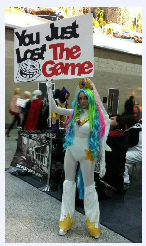 Photo of a person at a convention wearing colorful clothing and holding a sign reading 'You just lost The Game' with a trollface icon.