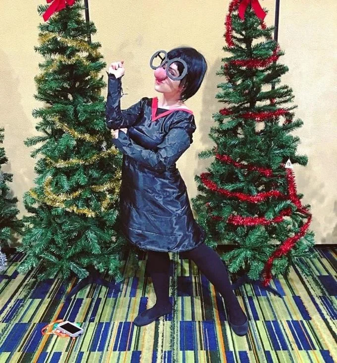The Incredibles — Cosplayer dressed as Edna Mode in a trash bag outfit between Christmas trees