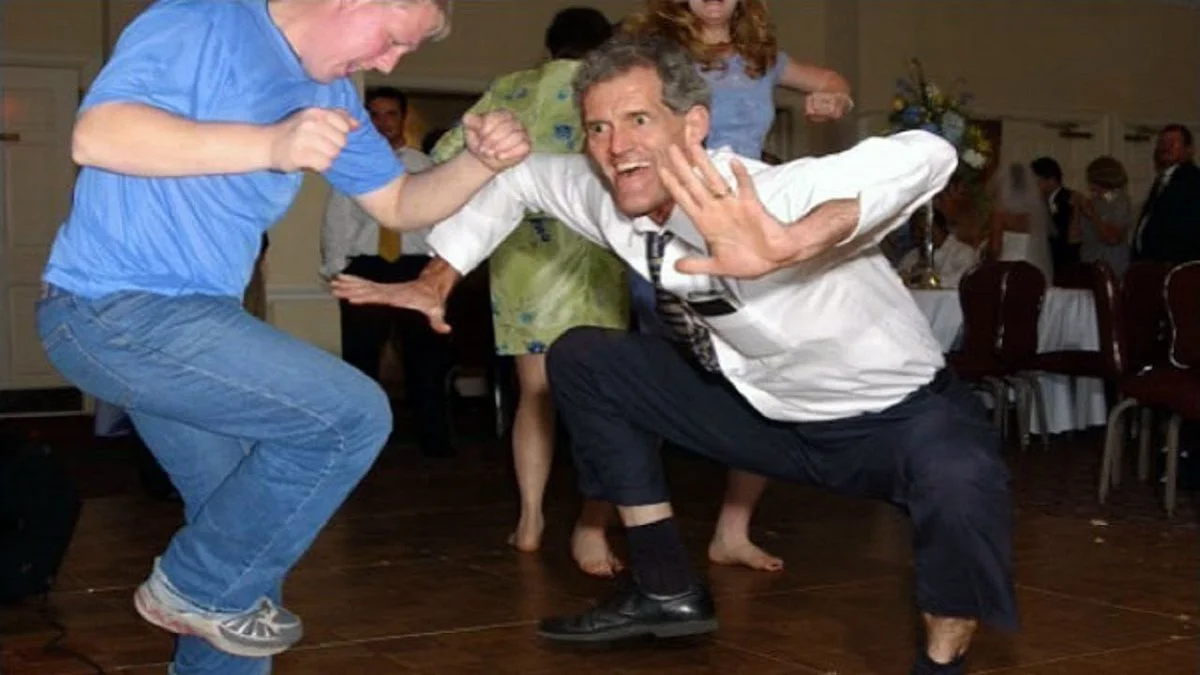The Jig — Barefoot man in dress shirt and tie crouching with exaggerated expression next to another man dancing at a wedding.