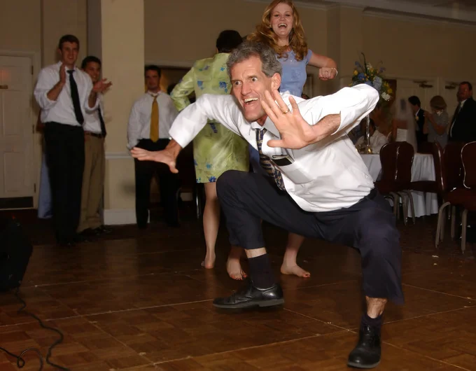 The Jig — Wider angle of the barefoot man crouching mid-dance at a wedding with onlookers in the background.