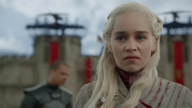 The Mad Queen — A production still of Daenerys Targaryen with platinum blonde braided hair and steely expression standing in front of castle banners.