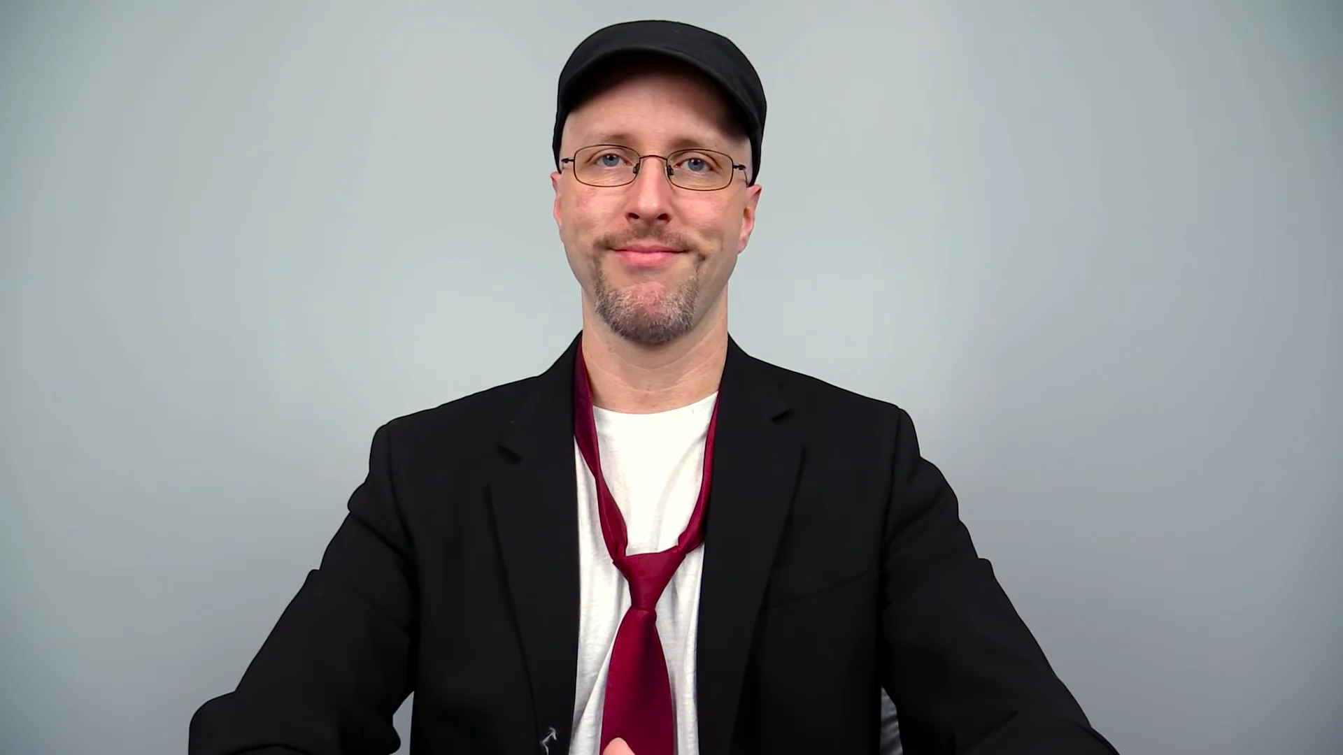 Nostalgia Critic Doug Walker smirking at the camera in cap, glasses, blazer, and red tie