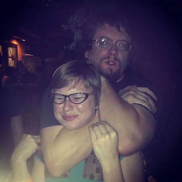 The Weak Should Fear the Strong — Bearded man in glasses with arm around smaller grimacing woman at a dark venue