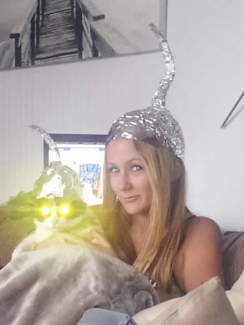 Woman and cat both wearing horned tin foil hats in a living room selfie.