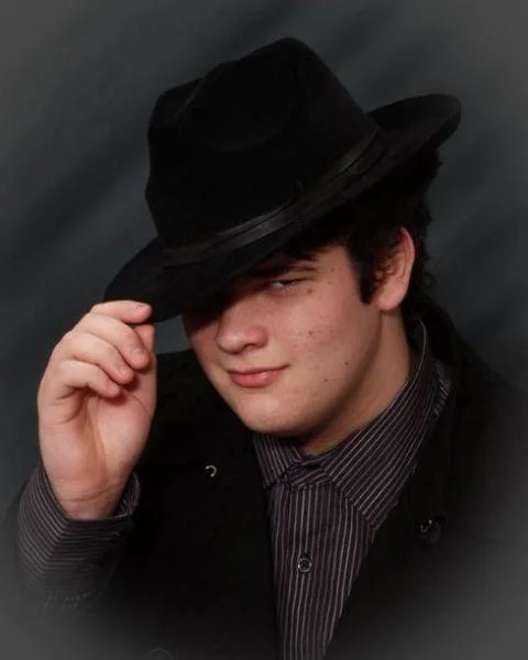*Tips Fedora* — Young man in pinstriped shirt tipping a black fedora in a dark studio portrait