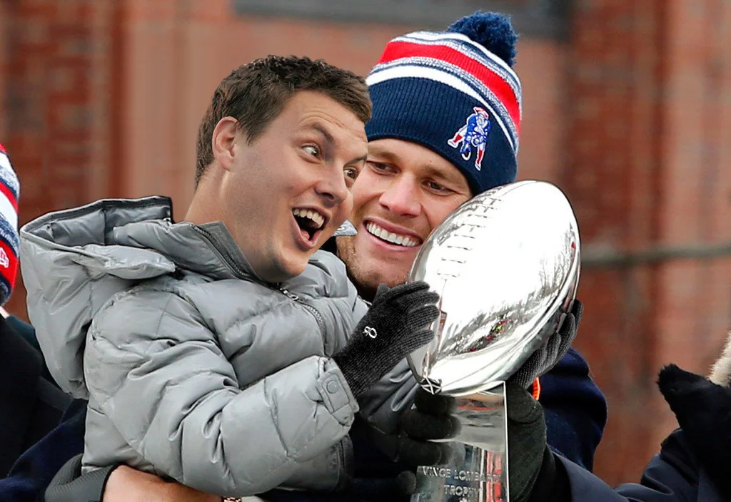Tom Brady wearing a Patriots beanie hugs a child while holding the Super Bowl trophy at a parade