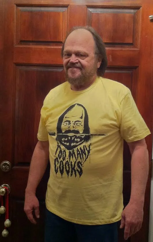 Man wearing a yellow Too Many Cooks T-shirt showing a bearded figure with a knife.
