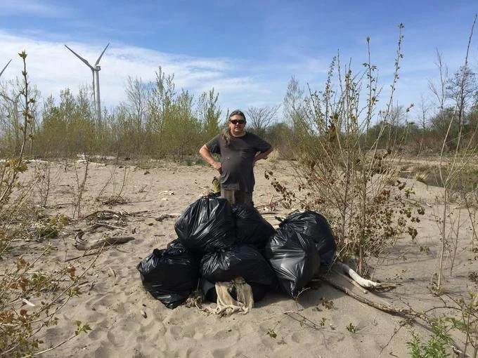 Trashtag — Person standing behind black trash bags on a sandy beach with a wind turbine in the background.