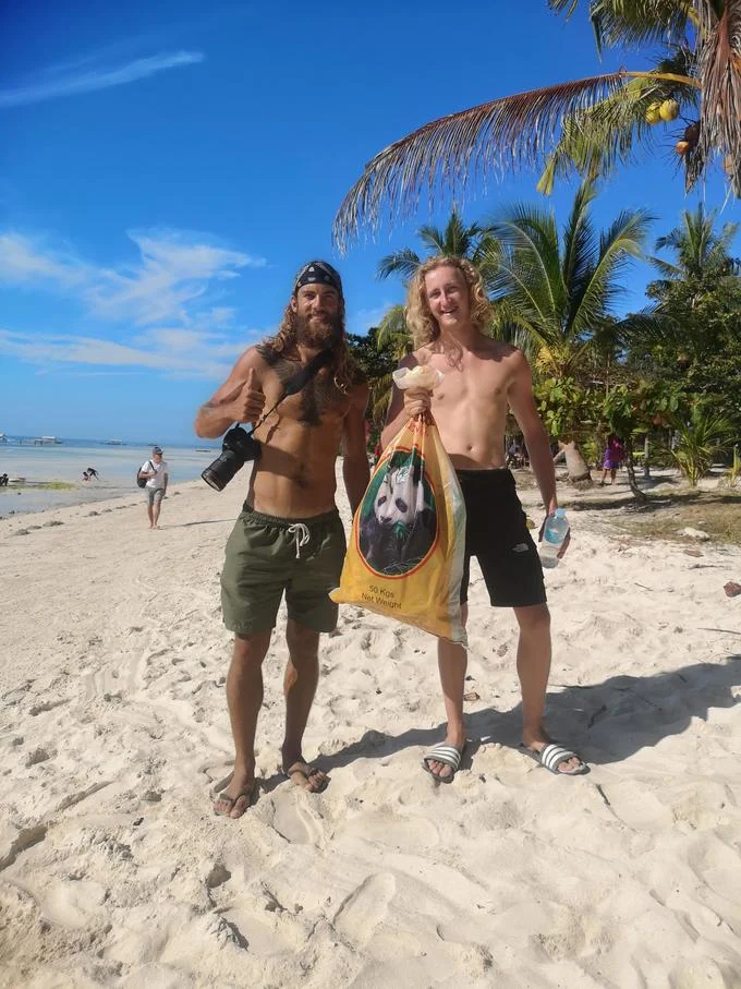 Trashtag — Two men on a tropical beach, one with a camera, the other holding a large bag of collected trash.