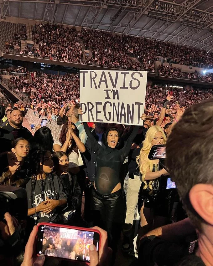 Travis I'm Pregnant Sign — Kourtney Kardashian in a sheer black top holding a 'Travis I'm pregnant' sign in a concert crowd.