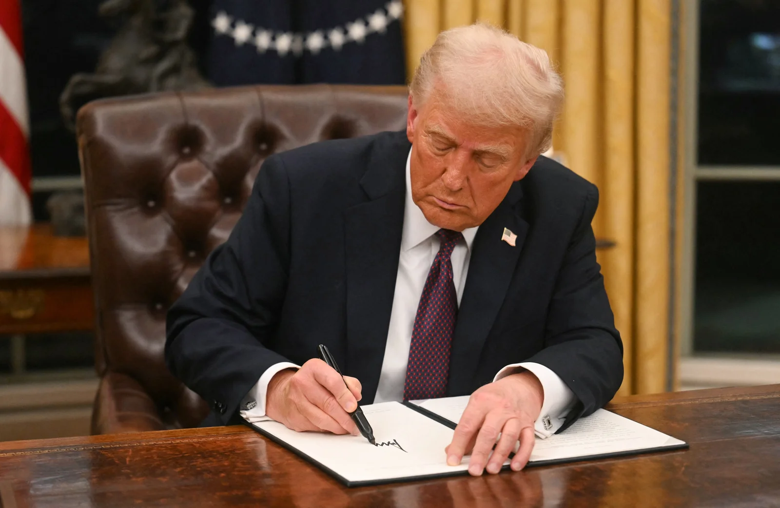 Trump and the executive order signature — Trump in formal business attire signing document at Oval Office presidential desk, clean source template photo.