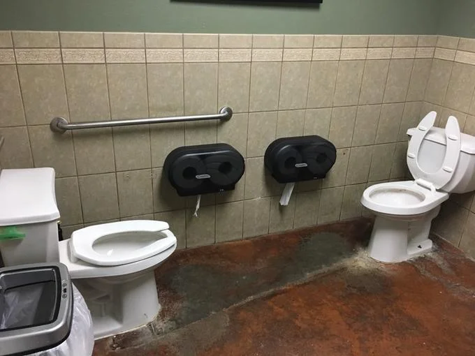Versus Pooping — Clear photograph of two facing toilets in a tiled public restroom with no partition