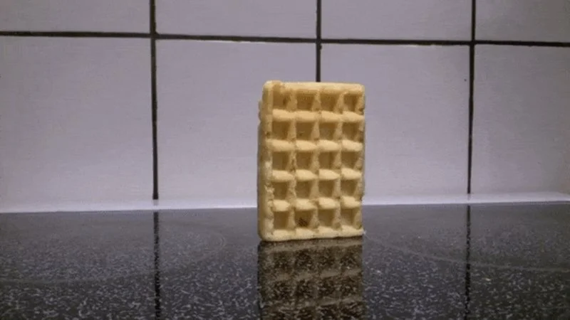 Waffle Falling Over Waffled — Golden-brown waffle standing upright on dark stovetop.