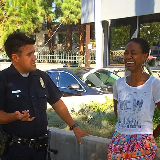 Walking While Black — Police officer speaking to a visibly upset Black woman on a sunny street next to a parked car.