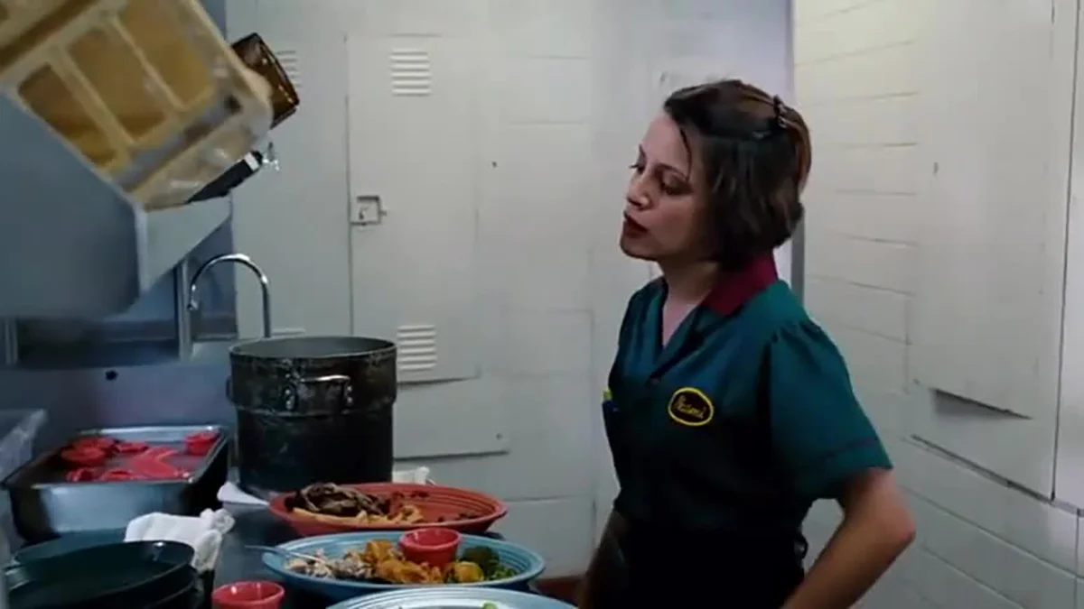 Wash The Fucking Dishes — Female restaurant worker in green uniform standing beside stacked dirty dishes in a kitchen