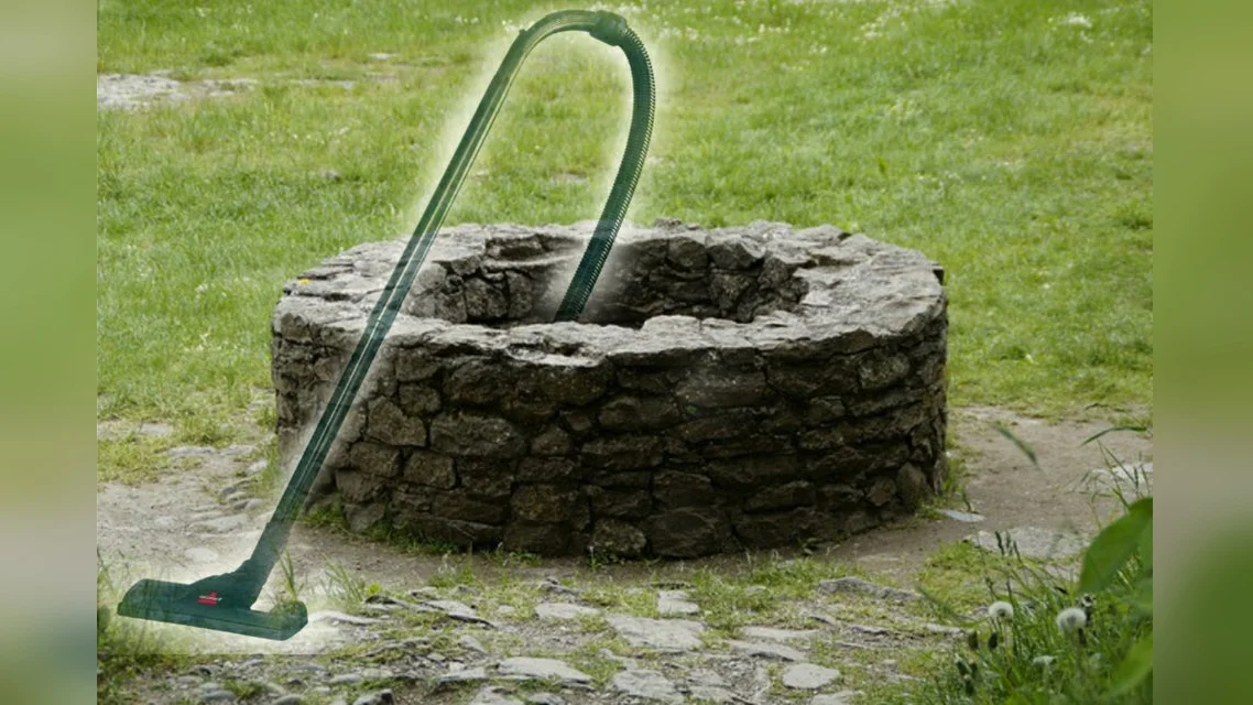 Well That Sucks — Stone wishing well with a green vacuum cleaner hose extending into it in a grassy field