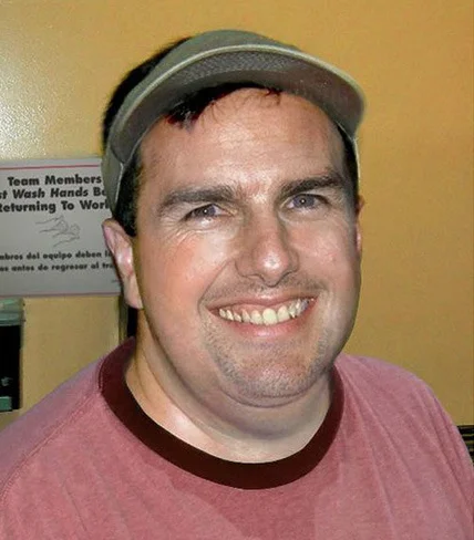 What Celebs Would Look Like Without Fame and Wealth Trend — Smiling man in a red t-shirt and gray cap resembling a celebrity lookalike in a workplace setting.