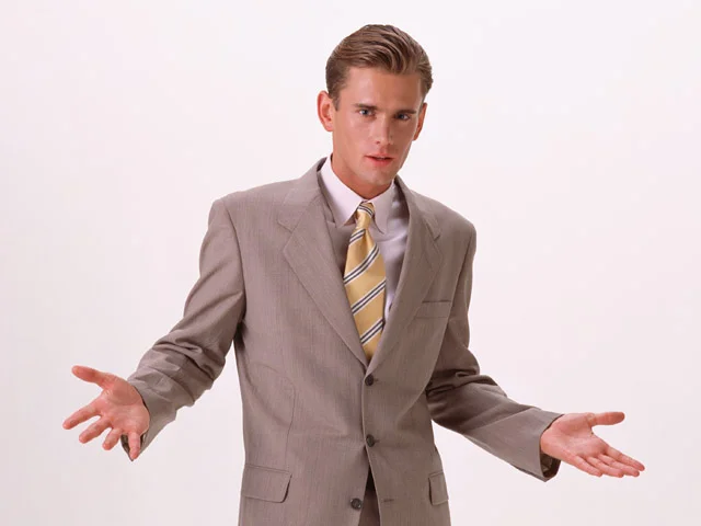 White Cis Male — Young white man in a tan suit shrugging with both hands out