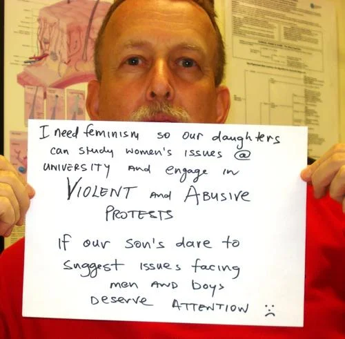 Who Needs Feminism — Person holding handwritten sign explaining feminism's importance for daughters to advocate on women's issues respectfully.