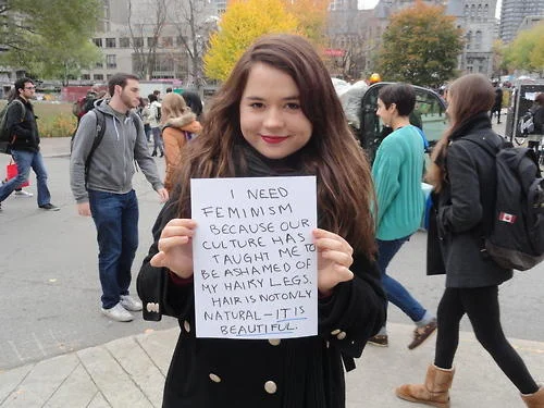 Who Needs Feminism — Young woman holding a handwritten sign explaining feminism's importance regarding body image and cultural pressures on women.