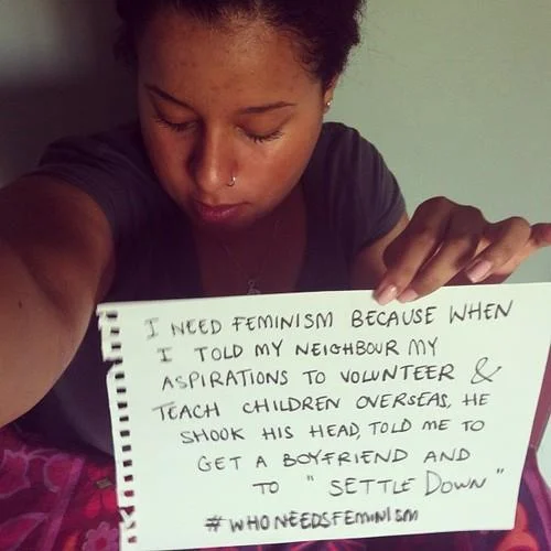 Who Needs Feminism — Close-up image of person holding handwritten sign about feminism and societal expectations regarding gender roles in volunteer work.